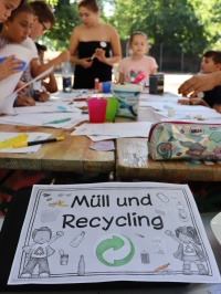 Sprachcamp 2022 Kinder arbeiten an einem Tisch mit Papiermaterialien, davor liegt ein Infoblatt zum Thema Müll und Recycling