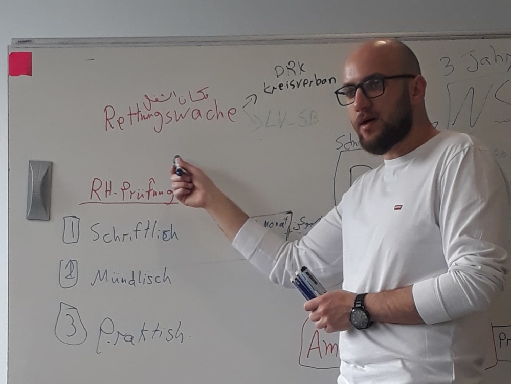 Unterrricht für Migrant*innen an der Rettungsdienstschule Saar Anas Alakkad steht vor Whiteboard, Blick Richtung Klassenraum, er zeigt auf die Tafel, Blick Richtung Klassenraum