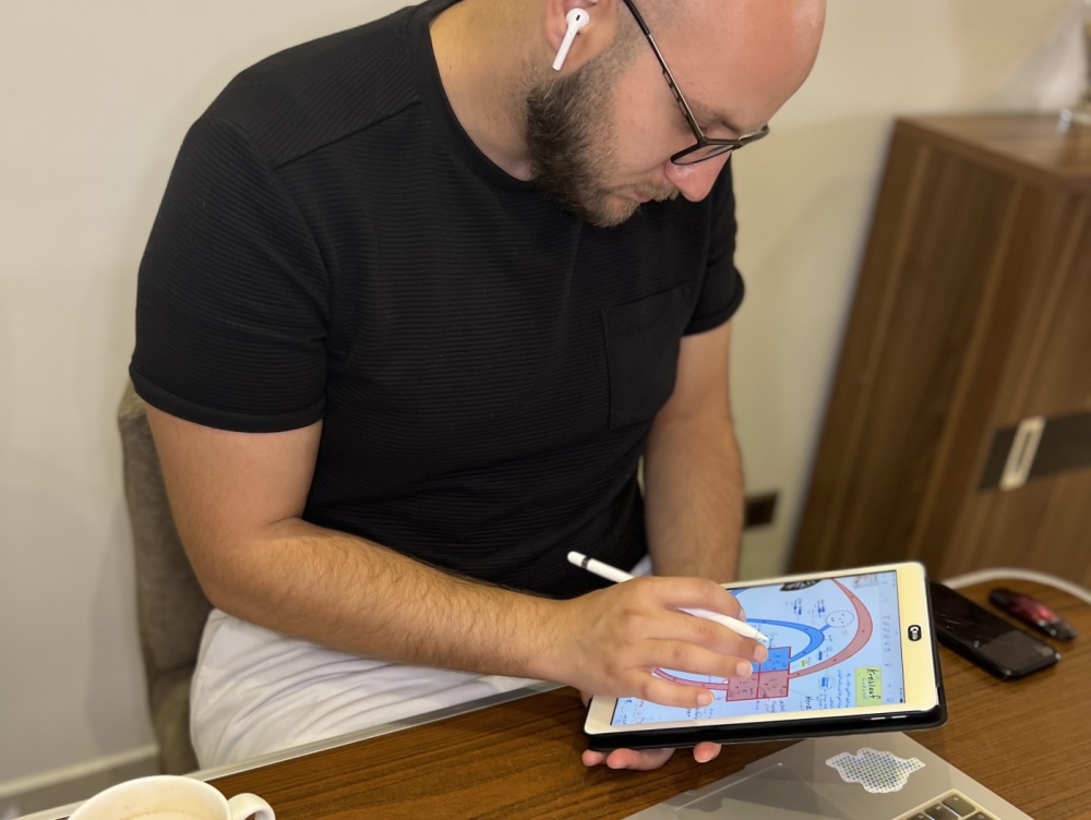 Online Unterricht Pontem Pro Herr Alakkad sitzt an Schreibtisch mit Laptop darauf und Tablet in der Hand. Auf dem Tisch befinden sich außerdem eine Maus und eine Kaffetasse.