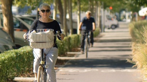 ältere Frau fährt auf Fahrrad Auf dem Foto ist ein Radweg zu sehen, der von Büschen und Gräsern umgeben ist. Im Vordergrund befindet sich eine Frau auf einem Fahrrad, weiter hinten ein Mann auf einem Fahrrad.
