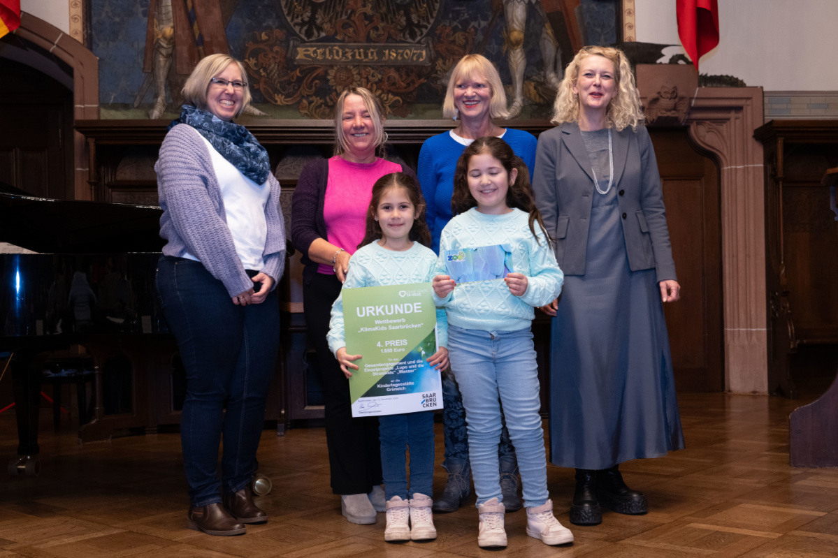 Platz 4 - Kita Grüneich Preisübergabe des vierten Platzes an die Kita Grüneich. Zu sehen sind zwei Kinder welche die Urkunde halten und 2 Betreuerinnen der Kita, sowie zwei Jurymitglieder.