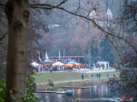 Blick durch Bäume auf die Stände des Weihnachtsgartens und Lichterglanz auf dem Wasser davor