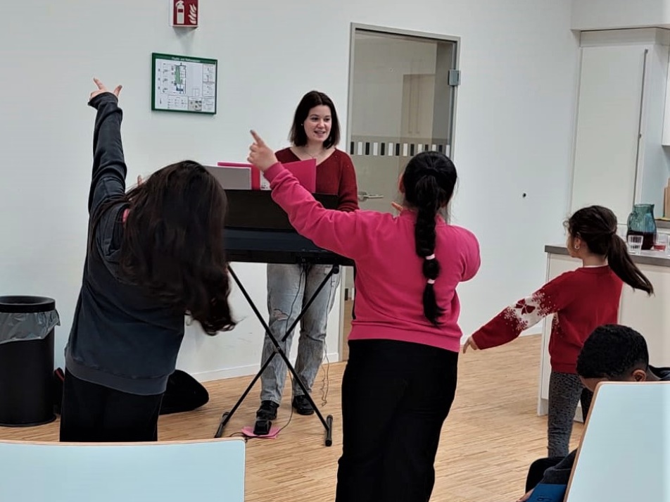 Kinder heben die Arme vor Notenständern und einer Frau Kinderchor in der Bildungswerkstatt