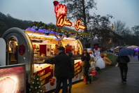 Ein Glühwein- und Secco-Stand mit leuchtendem Schild, davor Menschen, die Getränke bestellen oder trinken; im Hintergrund weihnachtliche Deko und ein großer aufblasbarer Schneemann.