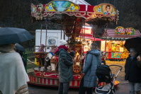 in kleines nostalgisches Karussell mit Kindern darauf, umgeben von Besucherinnen und Besuchern mit Regenschirmen; im Hintergrund leuchtende Essensstände.