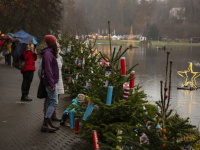 Menschen laufen am Seeufer entlang und betrachten eine Reihe kleiner Weihnachtsbäume, die mit buntem selbstgebasteltem Baumschmuck dekoriert sind; im Hintergrund leuchten Weihnachtsmarktstände und ein Stern auf dem Wasser.