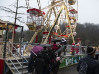 Menschen stehen in einer Schlange vor einem festlich geschmückten Riesenrad mit geschlossenen Gondeln, während ein Mitarbeiter Besucherinnen und Besucher einlässt.