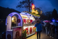 Ein glitzernder Wein- und Secco-Verkaufswagen leuchtet in buntem Licht, während Besucher mit Regenschirmen davor vorbeigehen.