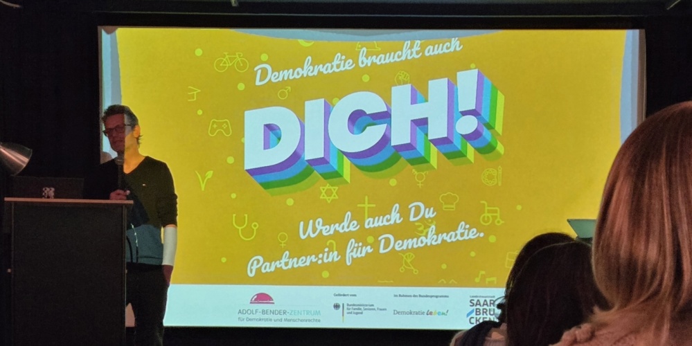 Demokratie leben Demokratiekonferenz 2025 Mann vor einer Leinwand mit der Aufschrift Demokratie braucht dich