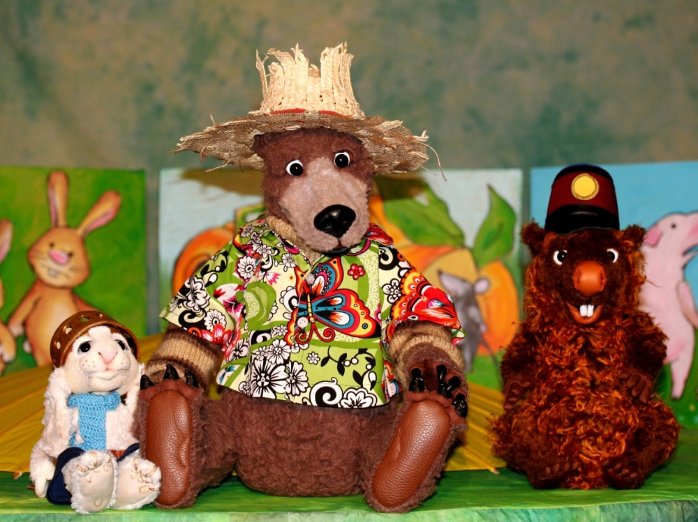 Szene aus "Hase und Holunderbär" Eine Bärenpuppe mit Strohhut und buntem Hemd, eine kleine weiße Hasenpuppe und ein Stoffmurmeltier mit Hut sitzen nebeneinander.