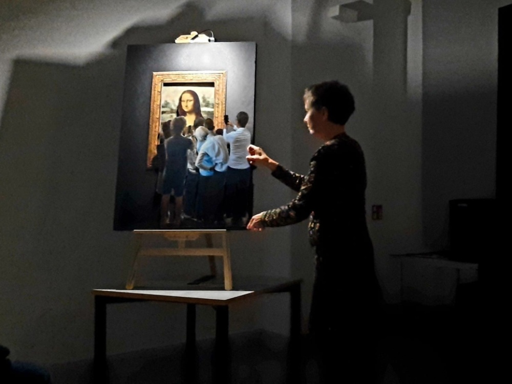 Szene aus "Das Lächeln der Mona Lisa?" Eine Frau steht neben einer Staffelei, auf welcher die Mona Lisa mit Besuchern und Besucherinnen abgebildet ist.