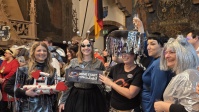Gruppe von kostümierten Erwachsenen steht dicht beieinander in einem historischen Saal mit Wandmalereien und Flaggen im Hintergrund. Die Personen tragen überwiegend futuristische oder silberfarbene Kostüme, einige mit glitzernden Perücken, auffälligen Bri