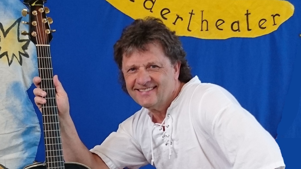 Eddi Zauberfinger Mann mit weißem Oberteil und roter Hose hat eine Gitarre in der Hand und sitz vor einem blauen Hintergrund