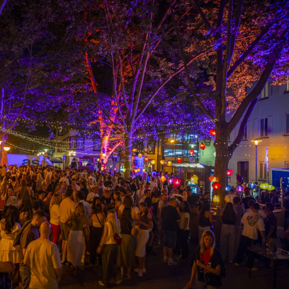 Eine belebte Altstadtstraße ist nachts mit bunten Lichtern, Lampions und vielen Besucherinnen und Besuchern gefüllt.